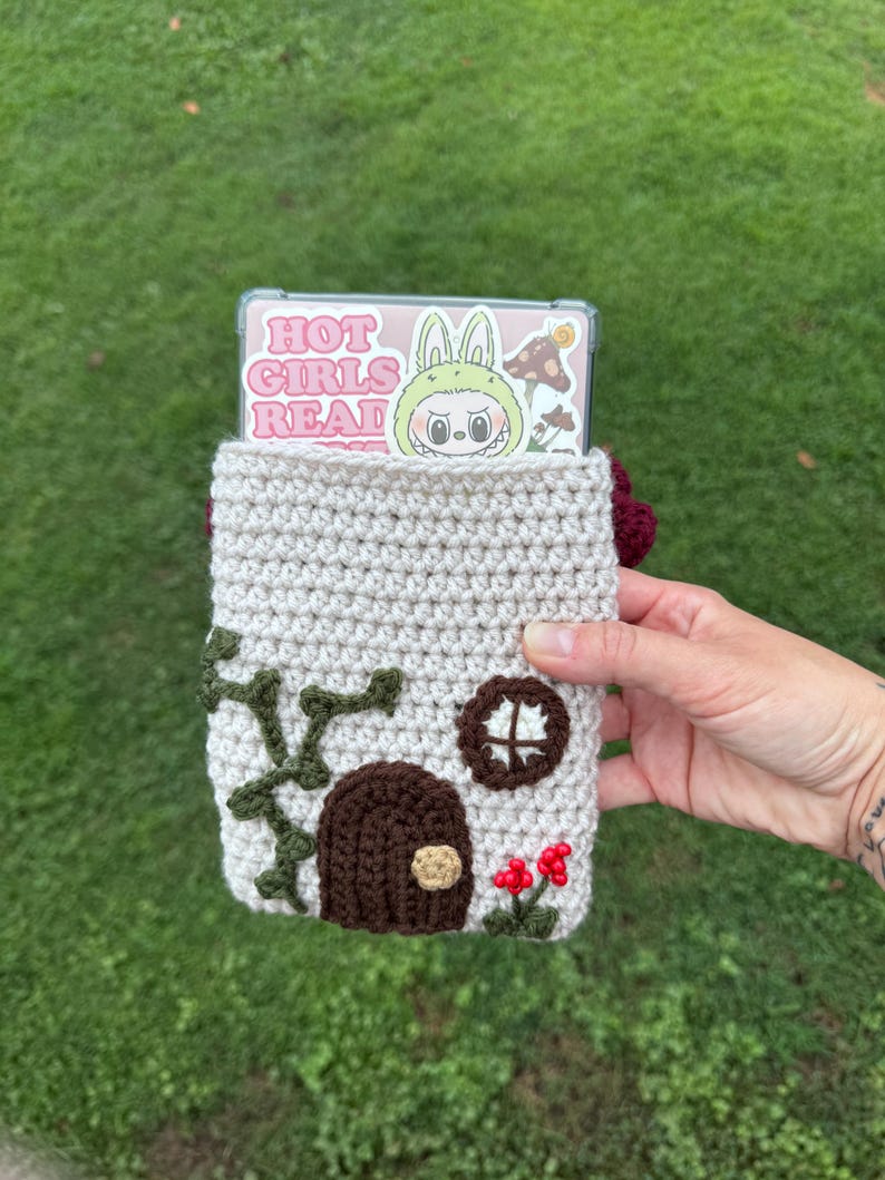 May include: A crocheted phone case in the shape of a house, with a brown door, green vines, and red flowers. The case holds a phone with a sticker that says "HOT GIRLS READ". The case is held against a green grass background.
