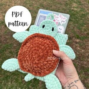 Op de afbeelding: Een gehaakt amigurumi-speelgoed in de vorm van een schildpad met een bruine schil en lichtgroene ledematen. Het speelgoed wordt vastgehouden in een hand met een tatoeage die "Ik hou van je" zegt.