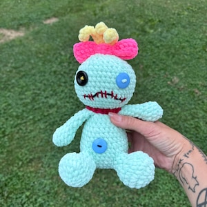 Pode incluir: Boneca de crochê azul claro feita à mão com um laço rosa, detalhes amarelos e olhos de botão. A boneca tem uma gola vermelha e uma boca costurada. A boneca é segurada em um fundo de grama verde.