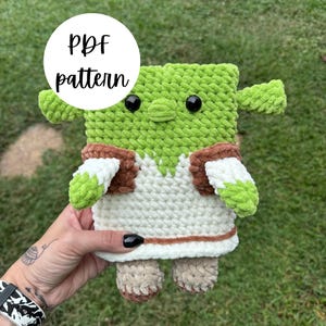 May include: A handmade green crochet plush toy with black eyes, small arms, and tan feet. The toy is wearing a white and brown outfit. A white circle with the words "PDF pattern" is in the background.