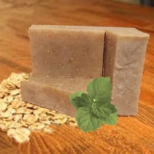 May include: A bar of handmade soap with oatmeal and mint. The soap is light brown and has a rough texture. It is sitting on a wooden surface with a pile of oatmeal in front of it.