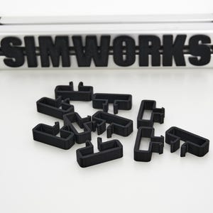 May include: A selection of black, T-shaped plastic connectors are arranged on a white surface. Above, a silver and black sign displays the word "SIMWORKS" in black lettering. The connectors are likely for a modular system.