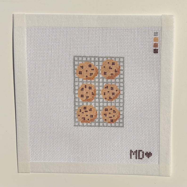 May include: A square needlepoint canvas featuring a grid of six light brown chocolate chip cookies on a gray cooling rack. The design includes a color palette guide in the top right corner and the letters MD followed by a heart in the bottom right.