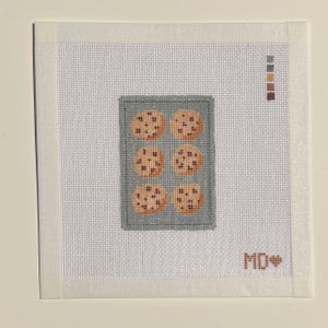 May include: A square needlepoint canvas featuring a gray rectangular baking sheet with six round tan cookies with dark brown chocolate chips. A vertical color palette of five thread swatches is in the top right, and the letters MD with a heart are in the bottom right.