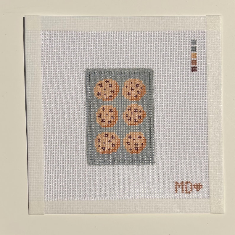 May include: A square needlepoint canvas featuring a gray rectangular baking sheet with six round tan cookies with dark brown chocolate chips. A vertical color palette of five thread swatches is in the top right, and the letters MD with a heart are in the bottom right.