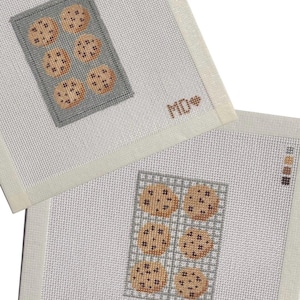 May include: Two square needlepoint canvases featuring cross-stitched designs of cookies on baking sheets. One canvas shows a gray baking sheet with six cookies and the letters MD with a heart. The other shows a grid-patterned sheet with six cookies.