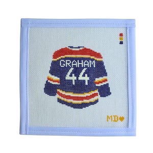 May include: Needlepoint hockey jersey with the name "GRAHAM" and the number "44" in white on a navy blue background. The jersey has red, yellow, and white accents. The needlepoint is framed in a light blue border.