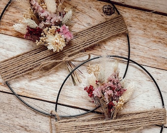 May include: Two black metal hoop wreaths decorated with dried flowers in pink, cream, and burgundy. The flowers are tied with twine. The wreaths are displayed on a light brown wooden surface.