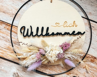 Op de afbeelding: Een rond houten bord met het woord "Willkommen" in zwarte letters en een klein hartje met het woord "lich" in gouden letters. Het bord is versierd met gedroogde bloemen en grassen in tinten wit, beige en paars. Het bord hangt aan een zwarte metalen ring.