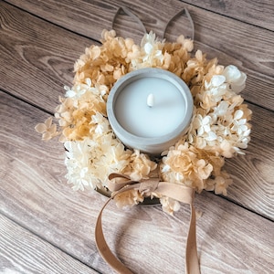 Könnte beinhalten: Eine weiße Kerze in einem Betonhalter, umgeben von einem Kranz aus getrockneten Blumen. Der Kranz ist mit einem braunen Band gebunden, auf dem "The Ostern" steht.