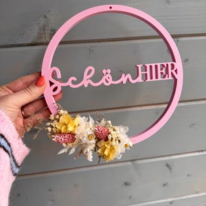 Puede incluir: Un letrero circular de madera rosa con las palabras alemanas "Schön Hier" y un arreglo floral. El letrero está sostenido por una persona. Las flores son amarillas, blancas y rosas.