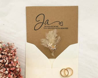 May include: A brown card with the word "Ja" written in cursive, followed by the text "Ein kleines Wort, das grosses Glück bedeutet."  The card is decorated with a small bouquet of dried flowers and is placed inside a white envelope with two gold rings.