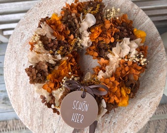 Op de afbeelding: Een ronde krans samengesteld uit gedroogde bloemen in bruine, oranje en crèmetinten. Een rond, lichtbruin acryllabel is bevestigd met een bruin lint, voorzien van de witte tekst "SCHÖN HIER" in een eenvoudig, schreefloos lettertype.