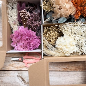 Könnte beinhalten: Ein Set getrockneter Blumen in einer Kartonbox. Die Box ist in drei Abschnitte unterteilt, die jeweils eine andere Blumenart enthalten. Die Blumen sind rosa, weiß und orange. In der Box befinden sich auch einige grüne Blätter und Stiele.