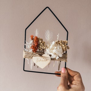 May include: A black metal house-shaped frame with dried flowers and a wooden heart that says "Zuhause" in white lettering.
