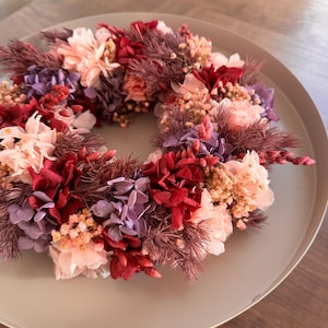May include: A floral wreath composed of various flowers in shades of red, pink, and purple, arranged in a circular shape. The wreath is placed on a round, light gray tray, creating a decorative display. The flowers appear to be dried or preserved.