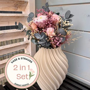 Könnte beinhalten: Eine beige, spiralförmig strukturierte Vase gefüllt mit einem Bouquet aus rosa und lila Blumen und dunkelgrünen Blättern. Die Vase steht auf einer schwarzen Oberfläche. Ein runder Aufkleber auf der Vase lautet "VASE & STRAUSS 2 in 1 Set DIREKT FERTIG."