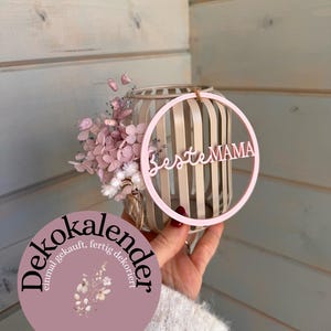 Può includere: Una composizione decorativa con un cerchio di legno rosa chiaro con la scritta "Beste MAMA" e un vaso cilindrico a forma di gabbia pieno di fiori rosa. È visibile anche un adesivo circolare con la scritta "Dekokalender" e "April".