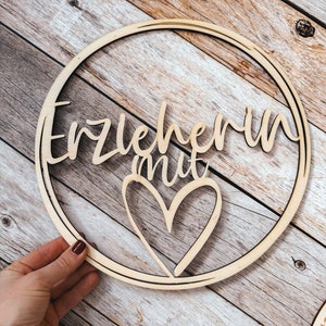 Può includere: Cerchio di legno con le parole "Erzieherin mit" e un cuore ritagliato. Il cerchio è su uno sfondo di legno.