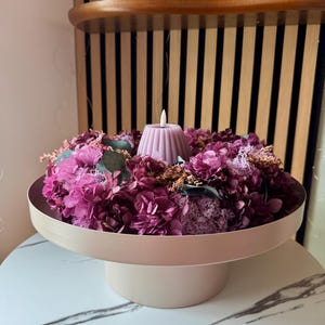 Könnte beinhalten: Dekoratives Centerpiece mit hellrosa Sockel, der ein kreisförmiges Tablett mit tiefrosa und lila Blumen und Grünzeug trägt. In der Mitte befindet sich eine hellrosa Kerze.
