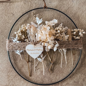 May include: A dried flower wreath with a heart-shaped wooden tag that says "Michaela & Matthias 01.09.2023" hanging from a black metal hoop.