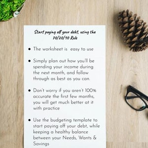 May include: A white sheet of paper with black text outlining the 70/20/10 rule for paying off debt. The text explains how to use the rule to budget and save money. The paper is on a wooden surface with a pine cone and a pair of glasses.
