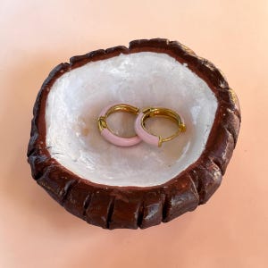 May include: A handmade jewelry dish resembling a coconut shell, with a white interior and a dark brown exterior. Two gold hoop earrings with pink enamel accents rest inside the dish. The earrings are small and delicate, adding a touch of elegance.