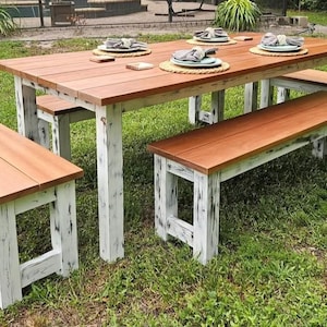 May include: A rustic outdoor dining set with a rectangular table and two benches. The tabletop and bench seats are stained a warm brown, contrasting with the distressed white legs. The set is placed on a grassy lawn.