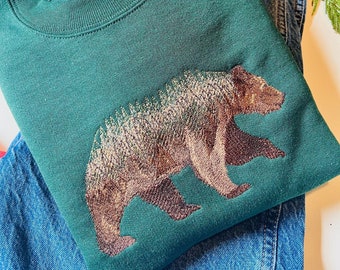May include: A dark green sweatshirt with a brown bear embroidered on the front. The bear is surrounded by a forest scene with trees and leaves. The sweatshirt is folded and lying on top of a pair of blue jeans.