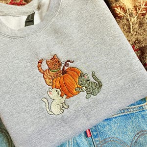 Puede incluir: Una sudadera gris claro con un diseño bordado de tres gatos y una calabaza. Los gatos son de color naranja, gris y blanco. La calabaza es naranja con un tallo verde. La sudadera está doblada sobre unos vaqueros azules.