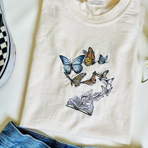 May include: A white t-shirt with an embroidered design of a book with butterflies flying out of it. The butterflies are in shades of blue, yellow, orange, and white.