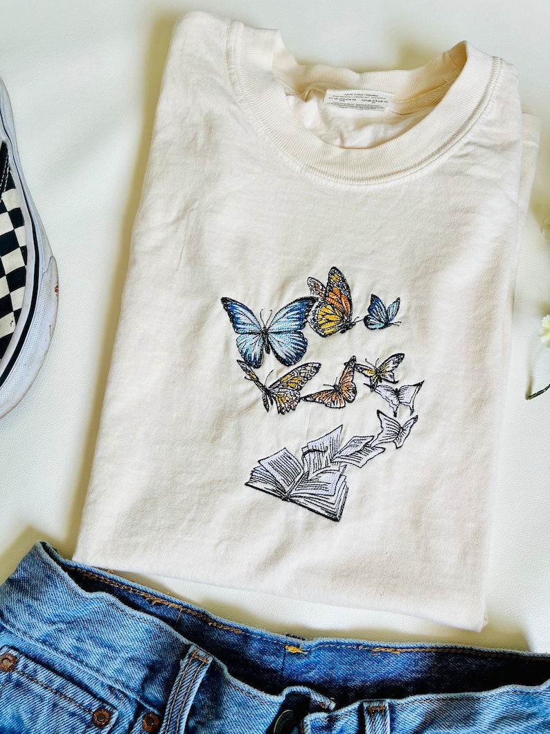 May include: A white t-shirt with an embroidered design of a book with butterflies flying out of it. The butterflies are in shades of blue, yellow, orange, and white.