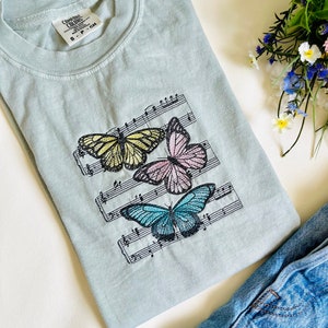 May include: A light blue t-shirt with a design of three butterflies on a musical staff. The butterflies are yellow, pink, and blue. The t-shirt has a tag that says "Comfort Colors" and "S - P - CH".