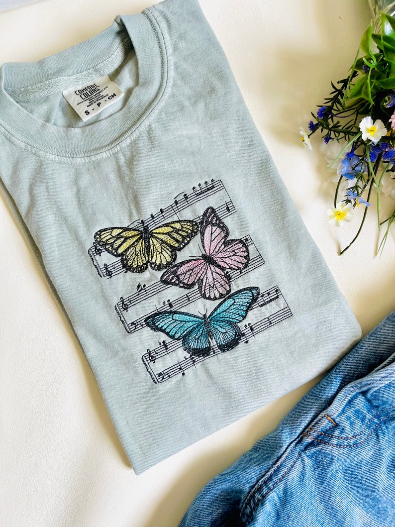 May include: A light blue t-shirt with a design of three butterflies on a musical staff. The butterflies are yellow, pink, and blue. The t-shirt has a tag that says "Comfort Colors" and "S - P - CH".