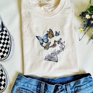 May include: A cream colored t-shirt with an embroidered design of butterflies flying around an open book. The t-shirt is folded and laid on a white surface with a pair of black and white checkered Vans shoes and blue jeans.