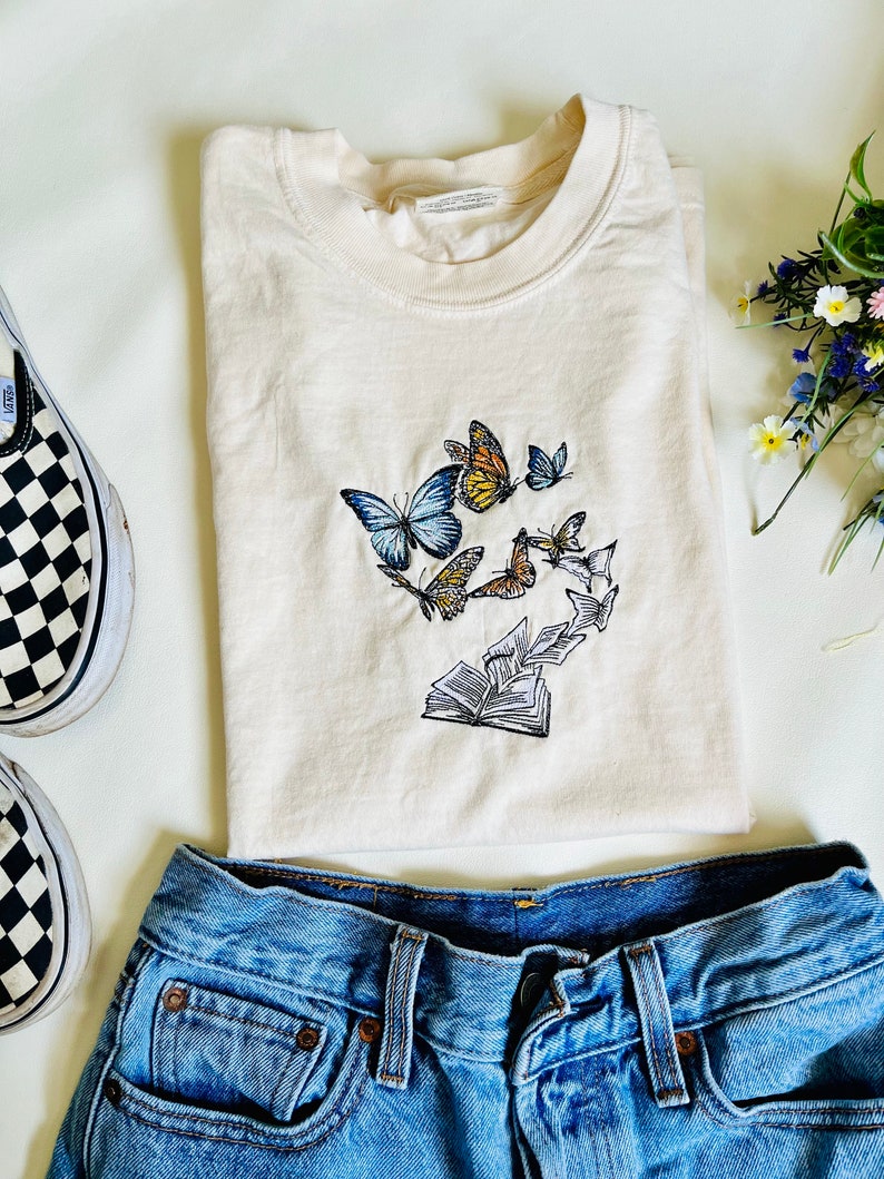 May include: A cream colored t-shirt with an embroidered design of butterflies flying around an open book. The t-shirt is folded and laid on a white surface with a pair of black and white checkered Vans shoes and blue jeans.