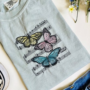 May include: A light blue t-shirt with a design of three butterflies on a musical staff. The butterflies are yellow, pink, and blue.