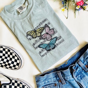 May include: A light blue t-shirt with a design of three butterflies on a musical staff. The butterflies are yellow, pink, and blue. The t-shirt is laid out on a white surface with a pair of black and white checkered Vans shoes and blue jeans.