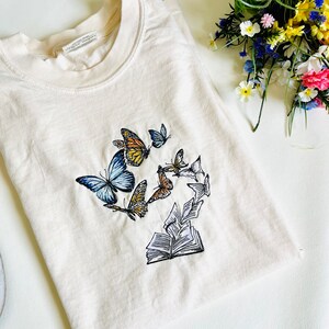May include: A cream-colored t-shirt with an embroidered design of a book with butterflies flying around it.