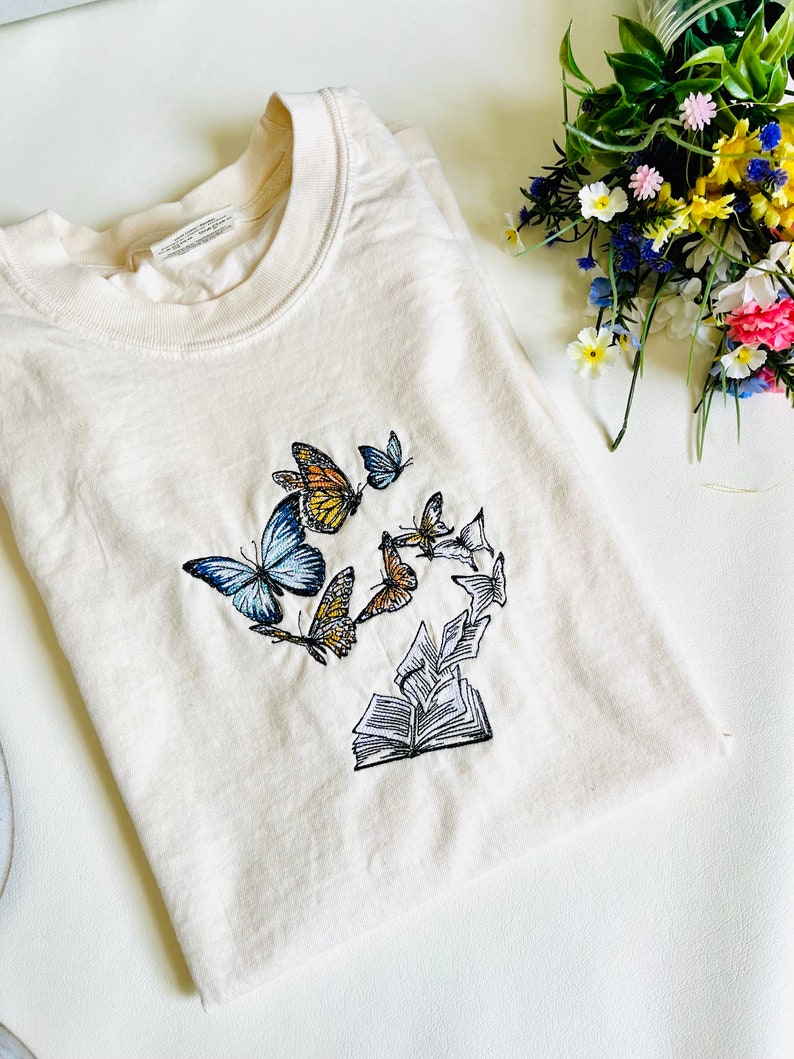 May include: A cream-colored t-shirt with an embroidered design of a book with butterflies flying around it.