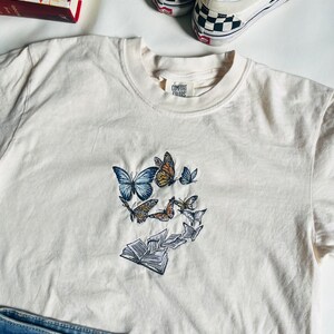 May include: A white t-shirt with a design of butterflies flying around a stack of books. The shirt has a tag that says "Comfort Colors".