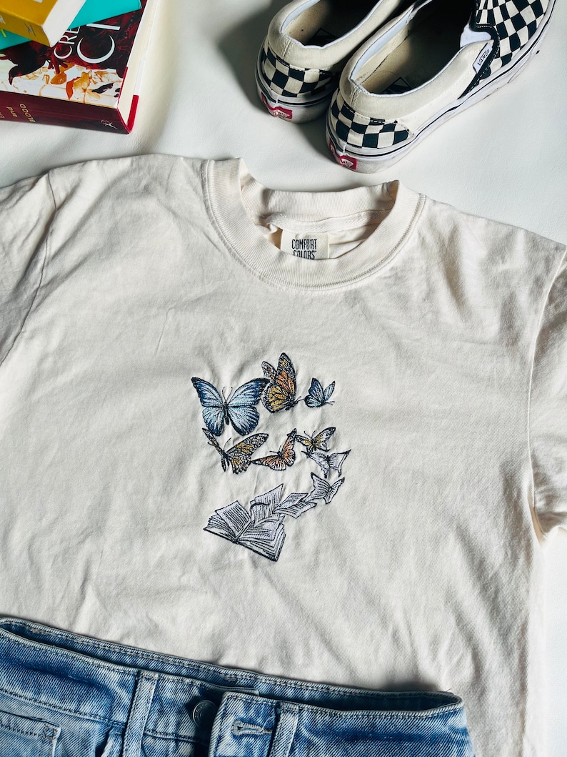 May include: A white t-shirt with a design of butterflies flying around a stack of books. The shirt has a tag that says "Comfort Colors".