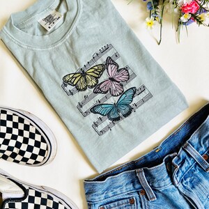 May include: A light blue t-shirt with a design of three butterflies on a musical staff. The butterflies are yellow, pink, and blue. The t-shirt is laid out on a white surface with a pair of black and white checkered Vans shoes and blue jeans.