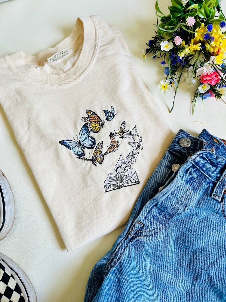 May include: A cream colored t-shirt with an embroidered design of a book with butterflies flying around it. The butterflies are in shades of blue, yellow, and orange.