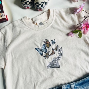 May include: A white t-shirt with an embroidered design of butterflies flying around an open book. The design is in shades of blue, brown, and yellow.