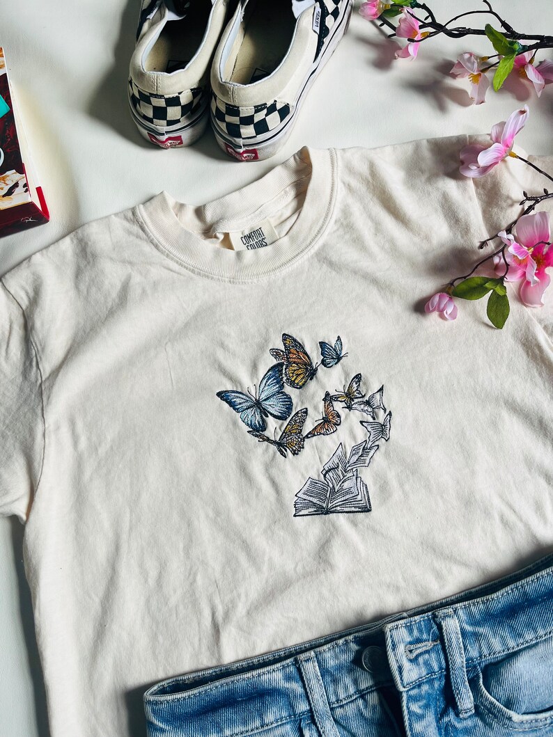 May include: A white t-shirt with an embroidered design of butterflies flying around an open book. The design is in shades of blue, brown, and yellow.