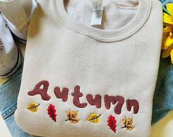 May include: A beige sweatshirt with the word "Autumn" embroidered in brown with small red, yellow, and orange leaves embroidered below.