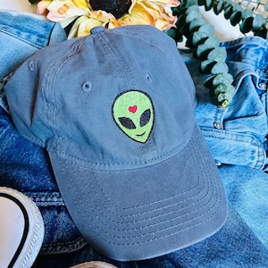 Puede incluir: Gorra de béisbol gris con un diseño bordado de una cara de alienígena verde con un corazón rojo. La gorra está sobre una chaqueta vaquera, con un girasol y vegetación de fondo. Tiene una visera curva y una correa de tela.