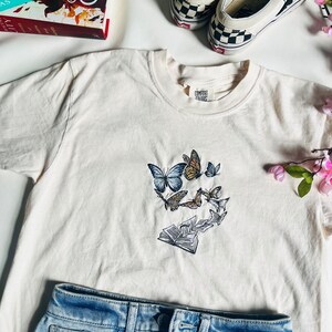 May include: A white t-shirt with an embroidered design of butterflies flying around an open book. The shirt has a label that says "Comfort Colors".