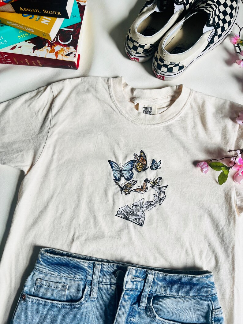 May include: A white t-shirt with an embroidered design of butterflies flying around an open book. The shirt has a label that says "Comfort Colors".